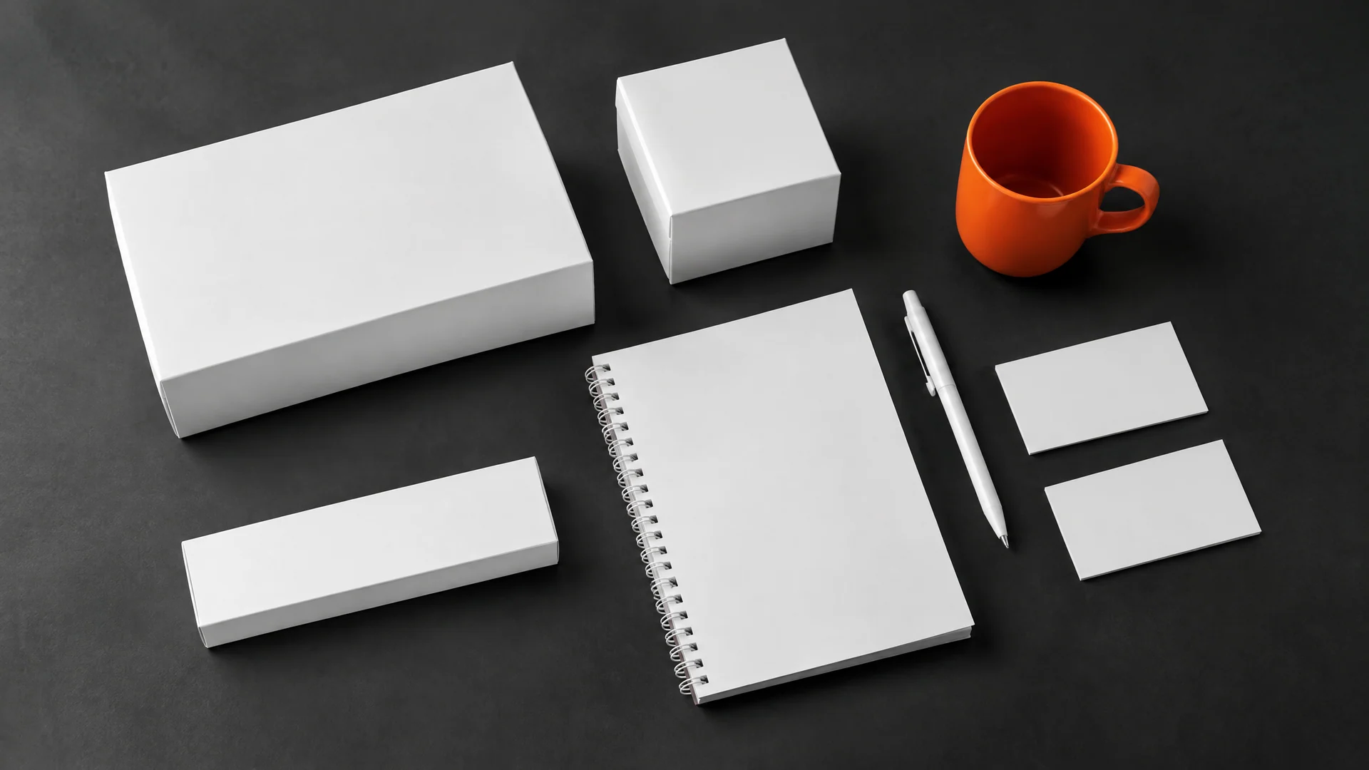 Blank white boxes, a spiral notebook, a pen, blank cards and an orange mug on a dark charcoal surface